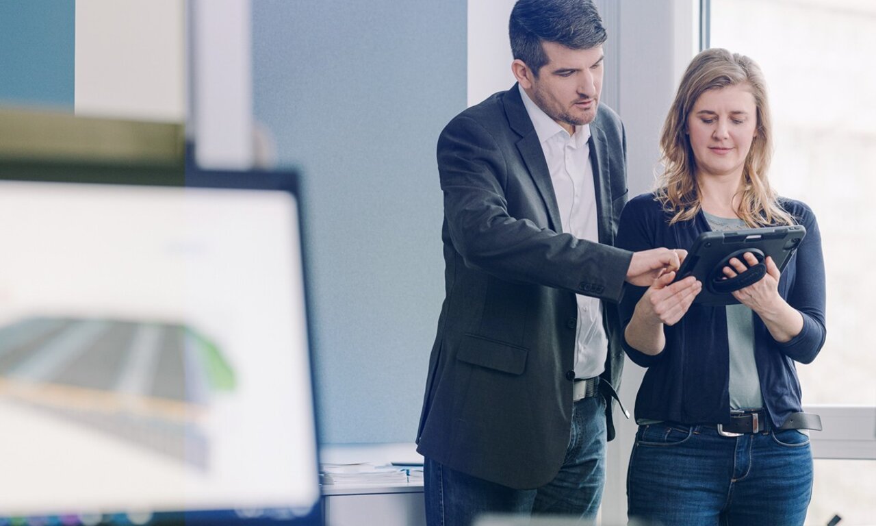 Zwei Mitarbeitende in einem Büro betrachten gemeinsam ein Tablet, während auf einem Bildschirm im Vordergrund ein digitales Modell zu sehen ist.